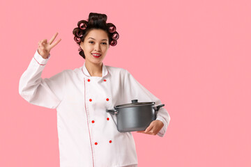 Female Asian chef with hair curlers and cooking pot showing victory gesture on pink background