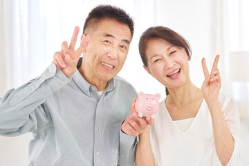 貯金箱を持ってピースサインするシニア夫婦　リビング