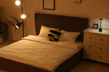 Interior of night bedroom with big bed, glowing lamps and commode near white brick wall