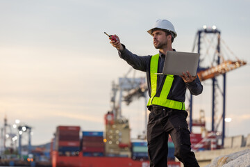 A male port engineer wearing a safety vest and helmet holds a laptop and talks on a radio at a busy...