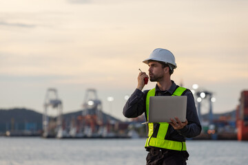 A male port engineer wearing a safety vest and helmet holds a laptop and talks on a radio at a busy shipping dock, overseeing cargo operations with cranes and containers in the background.