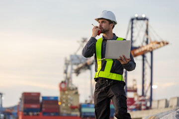 A male port engineer wearing a safety vest and helmet holds a laptop and talks on a radio at a busy shipping dock, overseeing cargo operations with cranes and containers in the background.