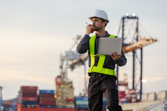 A male port engineer wearing a safety vest and helmet holds a laptop and talks on a radio at a busy shipping dock, overseeing cargo operations with cranes and containers in the background.
