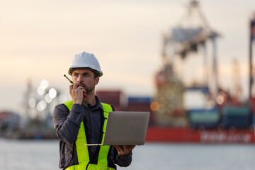 A port engineer wearing a safety vest and hard hat communicates via radio while holding a laptop, overseeing cargo operations at a busy shipping dock with cranes and containers in the background.