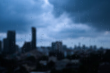 Raindrops on a window with a blurry city skyline in the background under a cloudy sky.