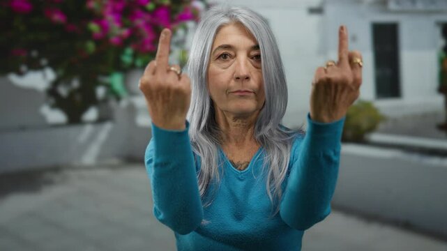Senior woman with grey hair making an offensive hand gesture on an outdoor street with blurred background during daytime.