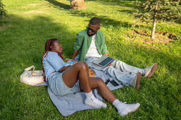 Relaxed African American couple study and work online in park on grass. Man researches exam details on device, woman assists engaging in discussion under trees in nature, remote work, e-learning