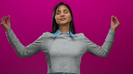 Young hispanic woman meditating with eyes closed against a vibrant pink background, embodying calm and serenity in a modern, minimalist setting.