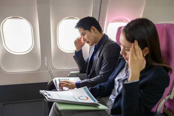 A man and a woman are sitting next to each other on a plane, looking at a laptop and a clipboard. They seem to be discussing something important, possibly related to their work or travel plans
