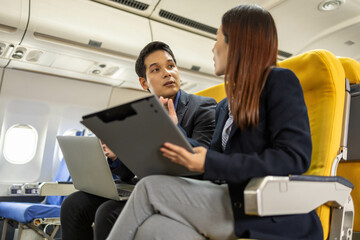 A man and a woman are sitting next to each other on a plane, looking at a laptop and a clipboard. They seem to be discussing something important, possibly related to their work or travel plans
