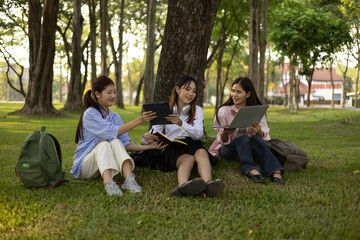 Three students sitting on the grass, one of them reading a book. The other two are looking at their laptops. Scene is relaxed and casual, as the women are enjoying their time together outdoors
