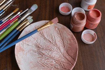mesa de trabajo de madera con pinturas, pinceles y plato de arcilla con textura de plantas