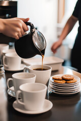 An office kitchen with coffee cups and plates on the table, a person is pouring coffee into a mug while another coworker grabs a cookie