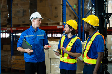 Logistics team of three male workers wearing safety helmets and vests engaged in discussion inside warehouse. One holding clipboard, others holding tablet and paper, with boxes stacked behind.