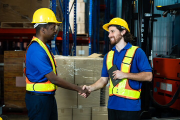 Two male warehouse workers in safety vests and helmets shake hands after completing successful logistics task. Industrial warehouse environment with boxes and forklift in background.