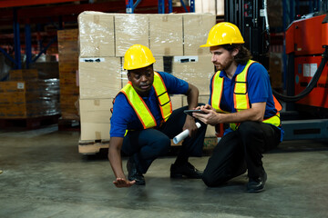 Two male warehouse workers wearing yellow helmets and reflective vests crouching on floor examining issue. One points at surface while other checks tablet in logistics storage facility.