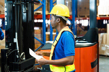 African male warehouse worker wearing safety helmet and vest holds checklist paper while monitoring...