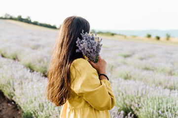woman in yellow dress stands in lavender field, gently holding bouquet of lavender flowers. She appears serene and contemplative, enjoying beauty and fragrance of blossoms around her.