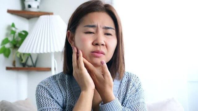Asian woman in discomfort, touching her cheek due to a throbbing toothache and swollen face, likely caused by serious oral problems such as cavities, gum disease, or dental abscess