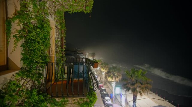 Static shot of a balcony covered with green plants, with street lights below and ocean waves crashing in the background at night, Letojanni, Taormina, Sicily, Italy (Italia)