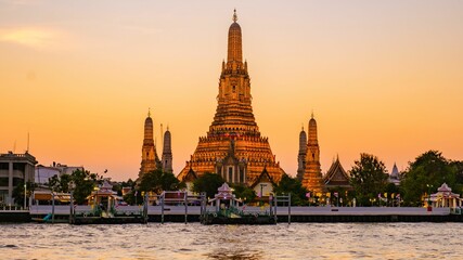 Fototapeta premium As dusk descends over Bangkok, Wat Arun radiates golden hues along the river. Visitors marvel at its stunning architecture, creating a tranquil atmosphere under the vibrant twilight sky.