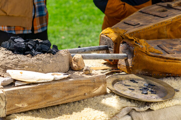 traditional outdoor blacksmithing or smelting setup featuring leather bellows, a clay furnace, charcoal, and hand tools. Likely part of a historical reenactment or craft demonstration