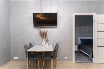 a large white dining table, grey chairs, a TV, grey walls, a white door. The design of a simple dining room or kitchen. The dining area.