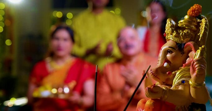 Focus on Lord Ganesha idol during Ganesh Chaturthi as Indian joint family with senior parents, son, daughter-in-law offer prayers with laddu plate and puja thali indoors in decorated festive home