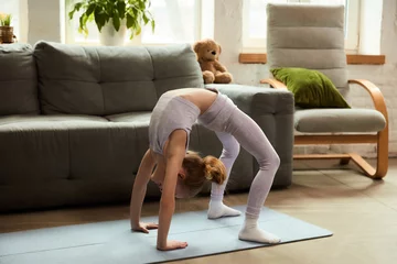 Fotobehang Flexible young gymnast holds challenging bridge pose yoga stretch on blue exercise mat, body forms arch, near grey sofa in living room. Concept of active child, lifestyle, hobby, sport at home. © Lustre Art Group 