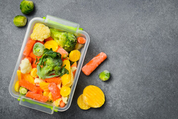 Frozen mix of vegetables in a plastic box. Preparation of frozen vegetables for the winter. On a gray stone background. Close-up.