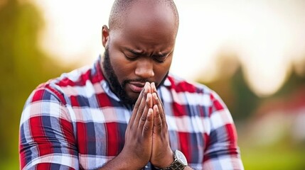 A man is praying in a park. He is wearing a red and white plaid shirt. The man is looking down and his hands are clasped together