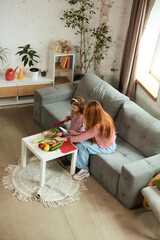 High angle view shows parent and child sitting together at small white table, carefully cutting...