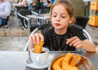 A young girl sits at a table in a lively cafe, carefully dipping a churro into a cup of chocolate