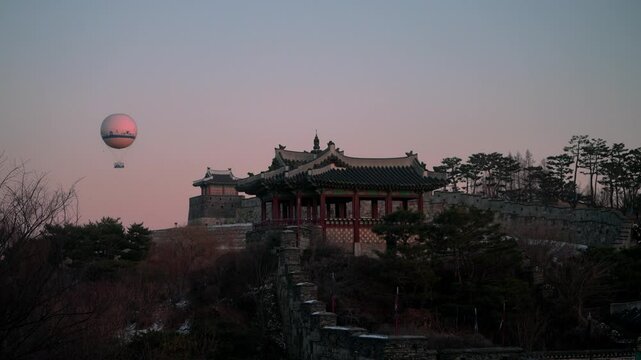Timelapse of a Korean traditional fortress at sunset. Hot air balloons rise and fall around the building, and flags bearing the Hanja 'command' and 'patrol' flutters in the wind.