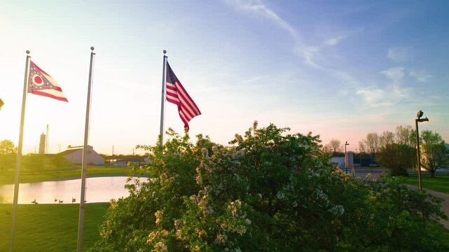 American and Ohio state flags waving at sunset beside industrial park and pond