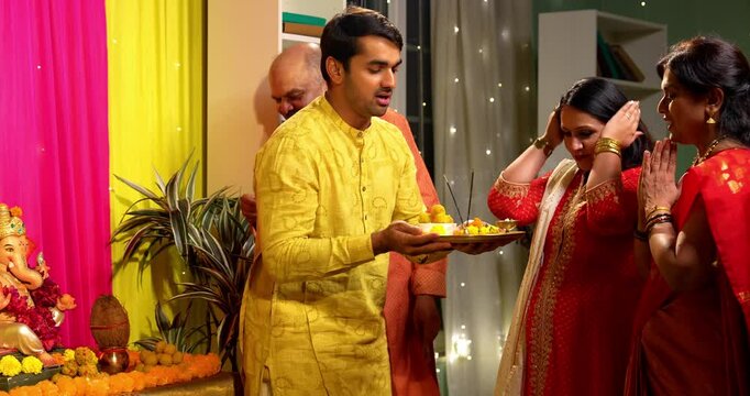 Distributing Ganesh festival prasad laddu, Indian family of senior parents young son with his wife dressed in traditional festive wear offering sweets performing Ganpati puja at home with devotion