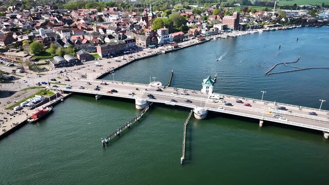 Aerial footage of the bascule bridge in Kappeln, Germany, with traffic and pedestrians, sailboats on the Schlei inlet and a busy harbor lined with old town buildings and caf&eacute;s.