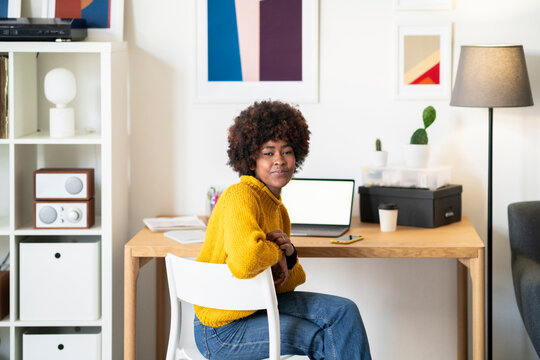 A young woman sits at her desk in a bright, modern home office. She is wearing a yellow sweater and jeans, and she is looking at the camera with a slight smile.