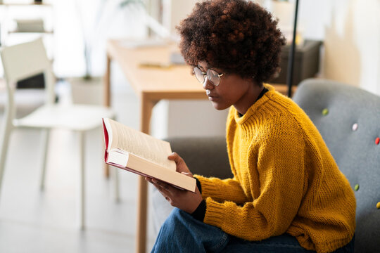 A young woman with glasses and an afro is reading a book. She is sitting in a chair in a bright, modern living room, enjoying a quiet moment with literature.
