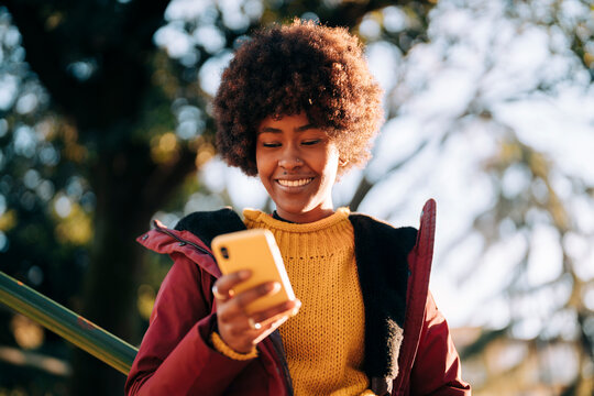 A young woman with an afro is smiling while looking at her phone. She is wearing a yellow sweater and a red jacket outdoors.