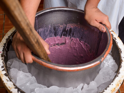 Hands churn purple ice cream in a metal bowl surrounded by ice.