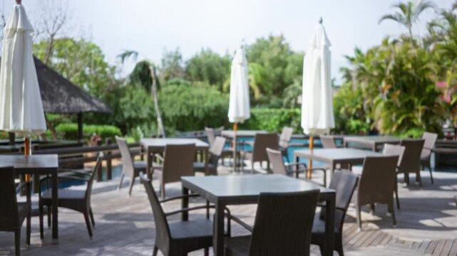 Blurred image of an outdoor tropical resort's luxury seating area with empty tables and chairs surrounded by lush greenery and parasols.