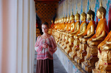 A woman stands in a corridor lined with golden Buddha statues draped in orange robes. The setting...
