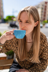Young woman with long blond hair, wearing a leopard print blouse, savors a hot drink from a light blue cup at a sidewalk cafe, enjoying the urban atmosphere