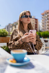 Young woman wearing sunglasses and holding smartphone while sitting at a table with a cup of coffee and a pastry, enjoying breakfast at an outdoor cafe