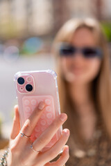 Close up of hands holding a smartphone with a pop it accessory and a transparent case, a young woman with sunglasses is blurred in the background, taking a selfie or recording a video