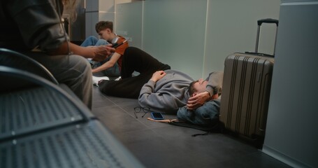 Crowded International Airport Terminal: Tired Man Sleeping on the Floor near Suitcase in Boarding Lounge of Airline Hub. Diverse Passengers Waiting for Delayed Flight. Tourism and Travelling. Handheld