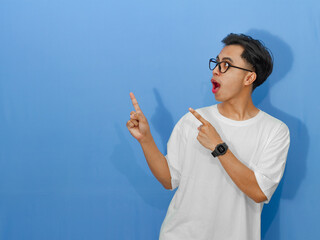 Excited young Asian man in a white t-shirt showing a funny surprise expression while pointing to the side.