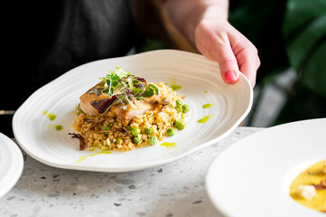 A hand holding a white plate with a gourmet dish featuring a perfectly cooked fish fillet atop grains and green peas, garnished with microgreens and drizzled with a vibrant green sauce