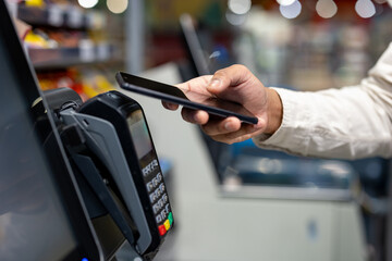 A person using a smartphone to make a contactless payment at a payment terminal in a store. It is a...